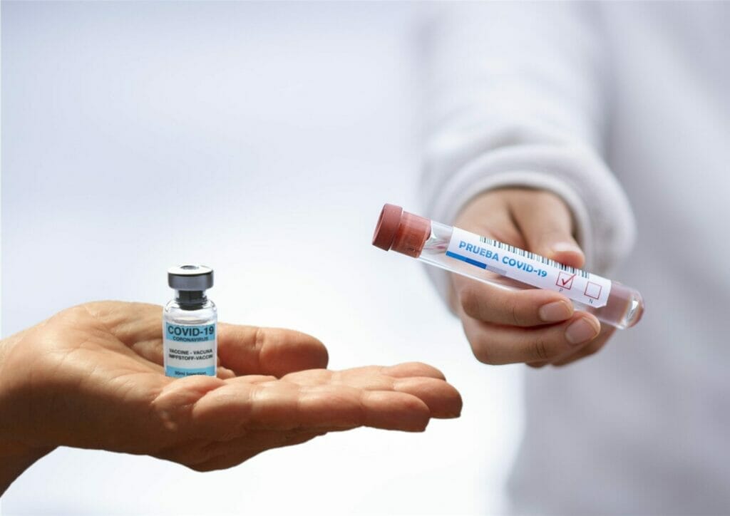 La imagen muestra una mano sostiendo un envase de vacuna contra covid-19 y otra mano sosteniendo un envase de prueba de covid-19.
