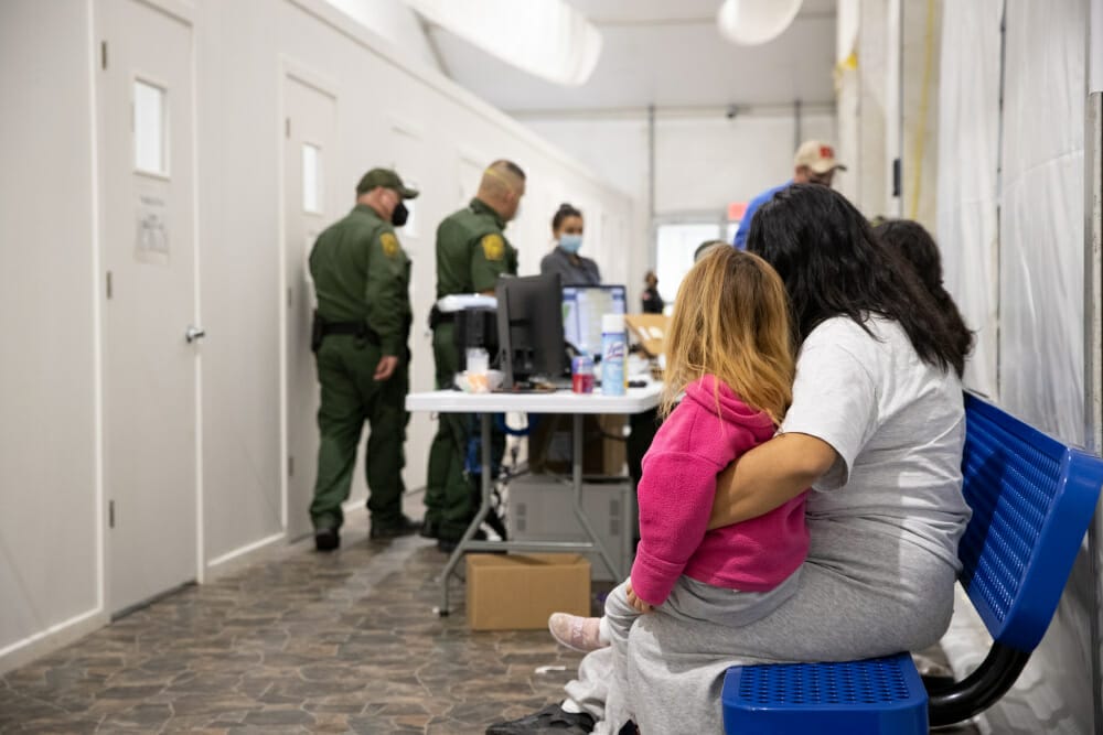 En la imagen se puede ver a una mujer adulta sosteniendo a una niña y detrás, a oficiales de la CBP - Este artículo habla sobre los niños en la frontera.