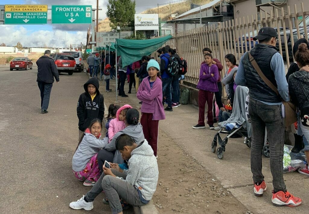 Este artículo habla sobre el Estatus de Protección Temporal para América Central. La imagen muestra a un grupo de migrantes esperando en la frontera sur de los EEUU.