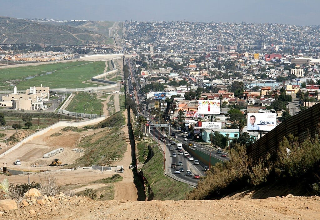 Este artículo habla sobre Quédate en México. La imagen muestra la frontera de México y Estados Unidos desde un punto elevado del camino fronterizo.