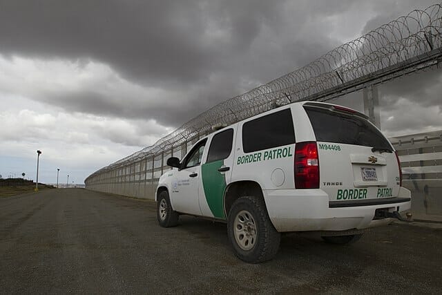 Este artículo habla sobre Quédate en México. La imagen muestra un vehículo de la patrulla de frontera junto a la reja fronteriza..