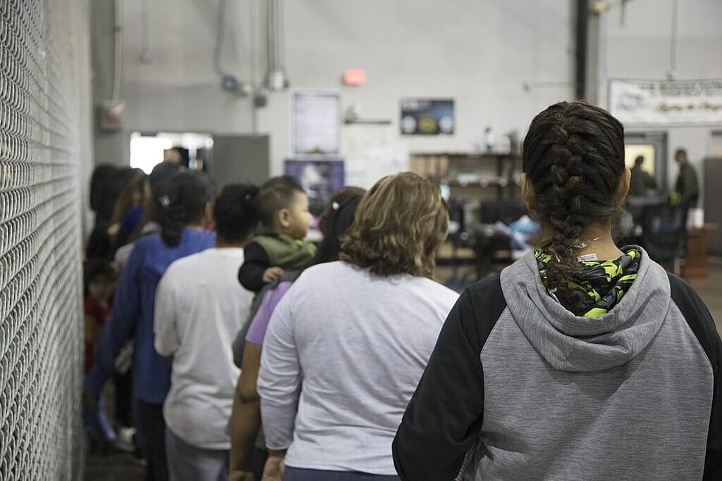 Este artículo habla sobre el acuerdo legal conseguido por personas detenidas en los centros de detención de inmigración. La imagen muestra un grupo de personas detenidas en uno de los centros vistas desde espaldas.