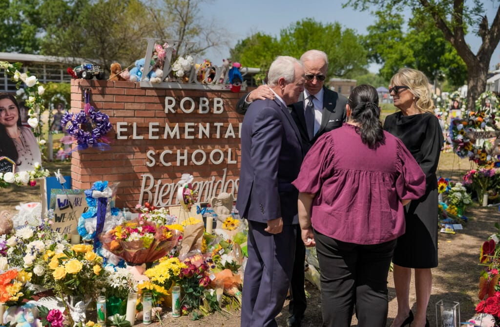 Este artículo habla sobre los comunicados emitidos por DHS y USCIS tras el tiroteo de Uvalde. La imagen muestra al presidente Joe Biden y otros individuos frente al cartel de la escuela primaria Robb, rodeada de flores en homenaje a las víctimas.