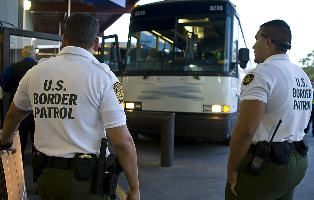 Este artículo habla sobre el plan de la administración Biden para lidiar con los altos números de cruces de frontera. La imagen muestra a dos oficiales de CBP a punto de registrar un bus.