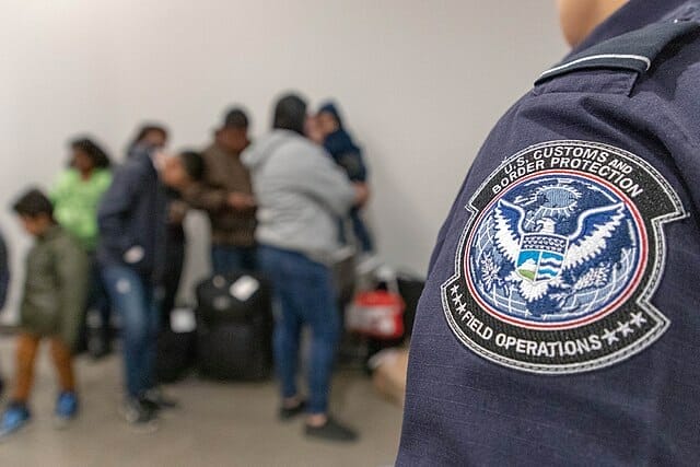 Este artículo habla sobre el fin del programa Quédate en México. La imagen muestra un grupo de solicitantes de asilo haciendo fila para ingresar a Estados Unidos. En primer plano se ve un hombro de una persona que viste uniforme de DHS.