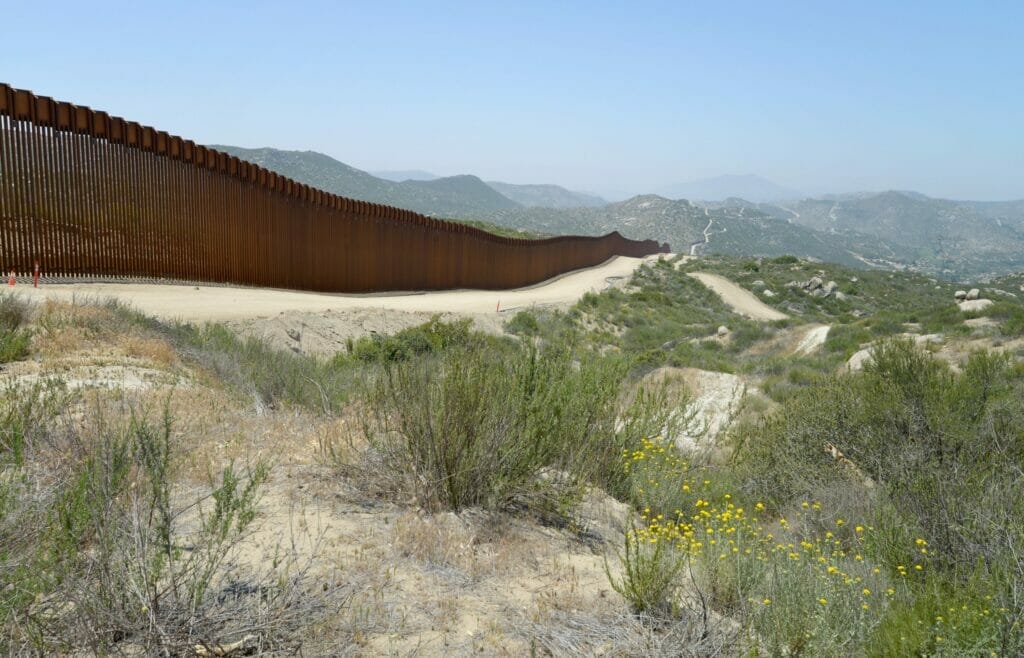 Este artículo habla sobre los cruces de migrantes en la frontera de baja California. La imagen muestra el muro fronterizo de California.