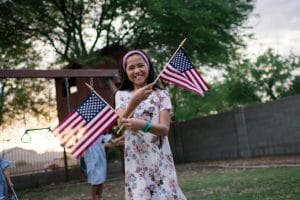 Este artículo habla sobre la ley "California ID's for All", que otorgará tarjetas de identificación estatal a residentes sin papeles. La imagen muestra una niña sonriendo. Sostiene dos banderines de Estados Unidos.