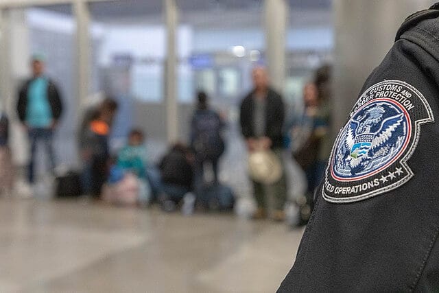 Oficial de CBP en puesto fronterizo y migrantes al fondo