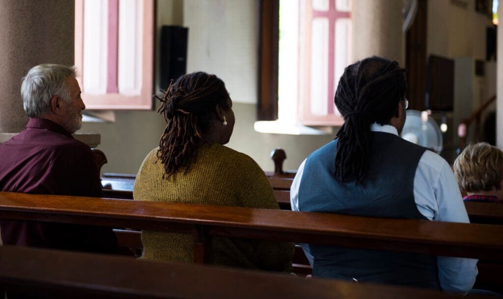 Tres personas de espaldas sentadas en el banco de una iglesia escuchando la misa. 