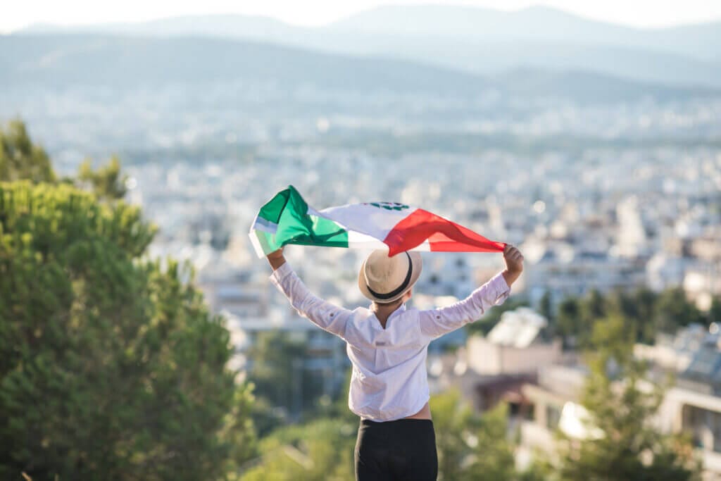 Niño sosteniendo la bandera de México con orgullo frente a un gran paisaje. 