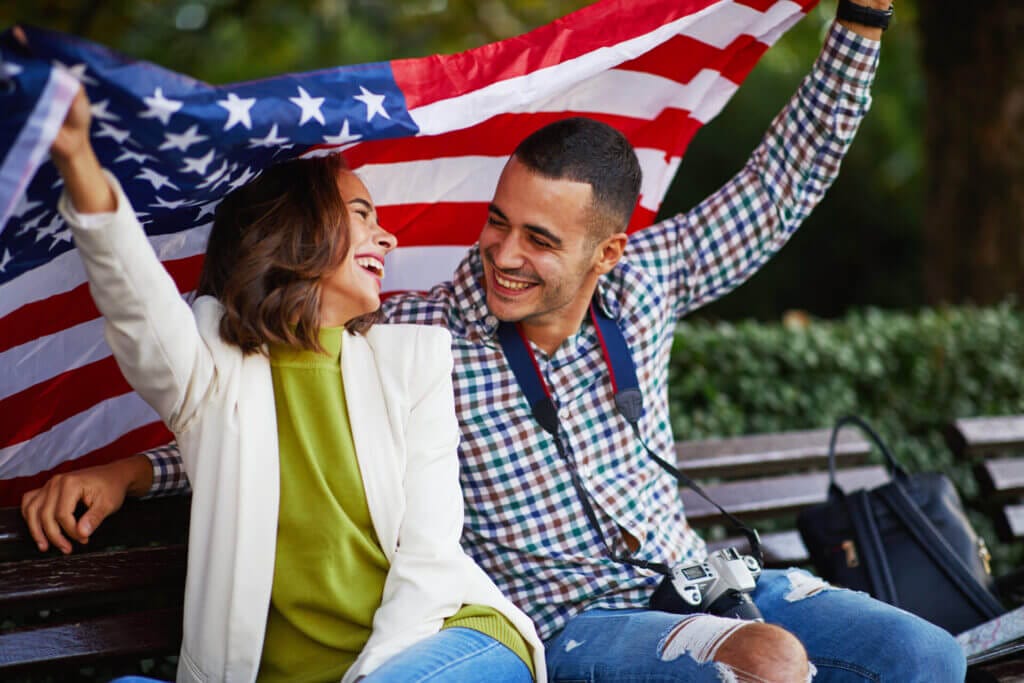 Pareja feliz en un banco de plaza con la bandera de EE.UU., contentos gracias a su visa de turista estados unidos