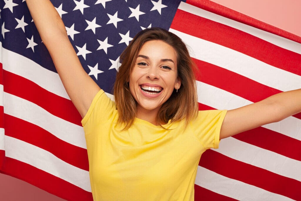 Mujer entusiasmada sonriendo con la bandera de EE.UU. en sus hombros tras obtener su visa