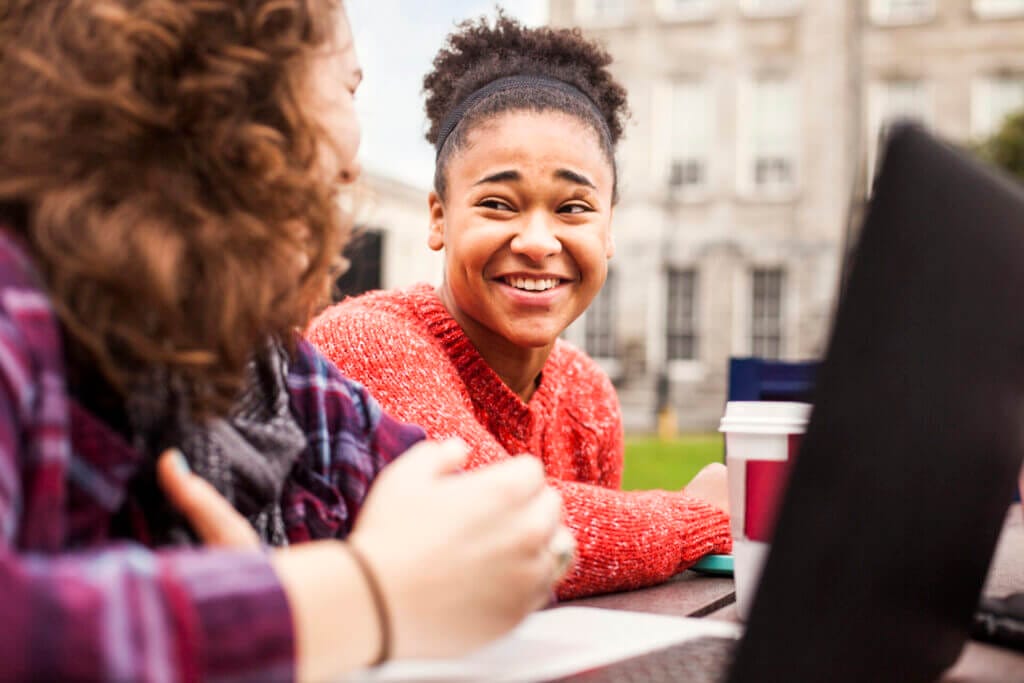 Estudiante en el patio del campus frente a una laptop riendo con una compañera