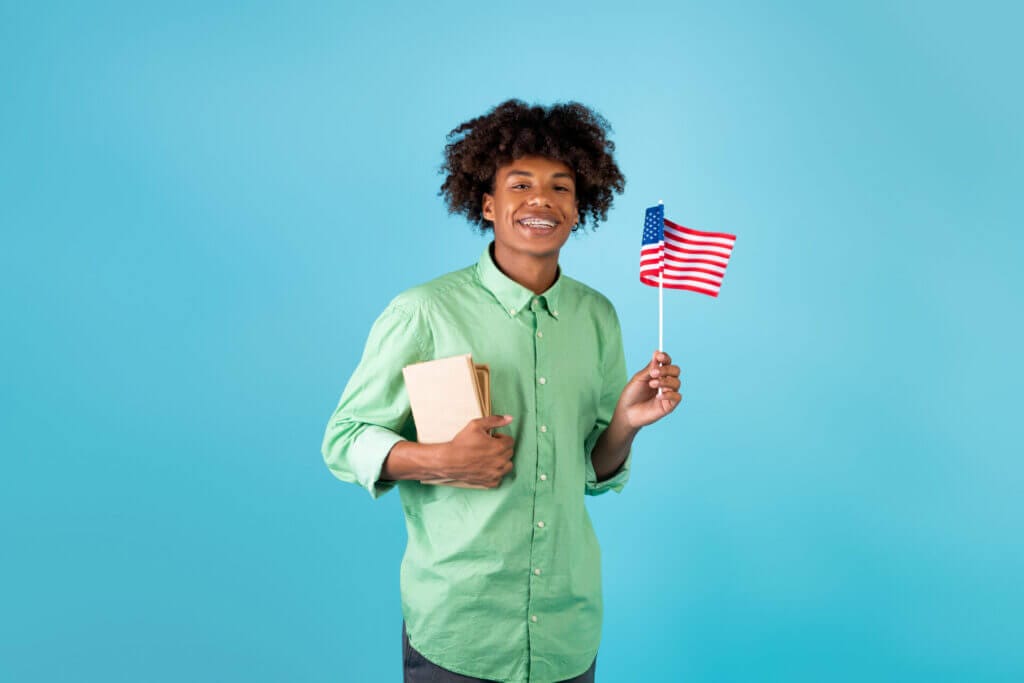 Estudiante de intercambio en Estados Unidos sonriendo a la camara con sus cuadernos y la bandera de USA