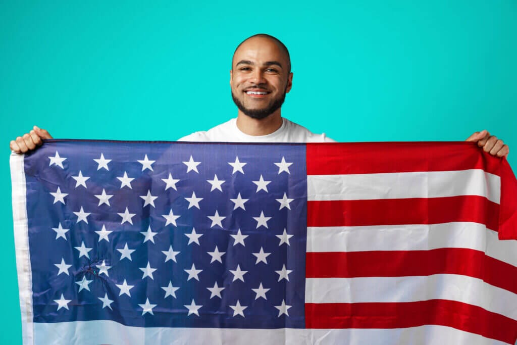 Retrato de un hombre migrante sonriendo mientras sostiene la bandera de USA