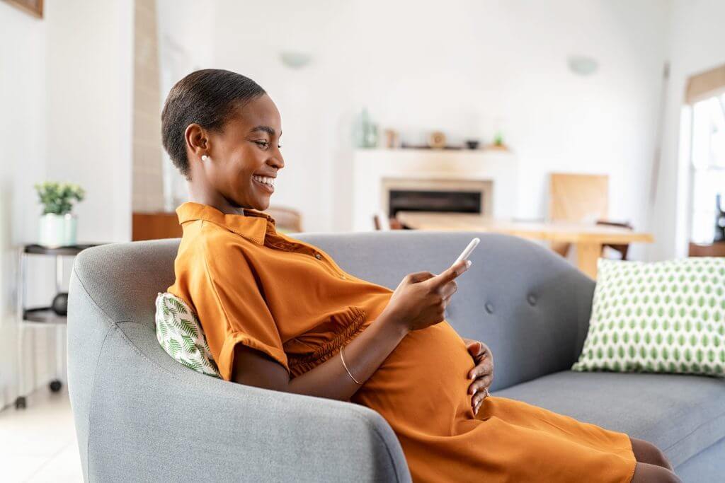 Mujer embarazada descansando en su sofá mientras sonríe al teléfono