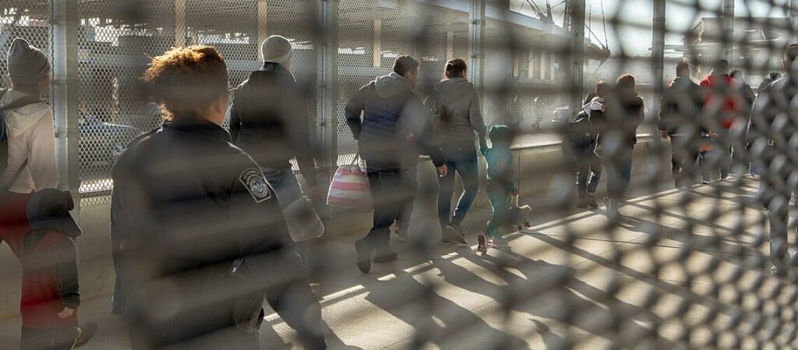 Este artículo habla sobre el fin del programa Quédate en México. La imagen muestra un grupo de solicitantes de asilo haciendo fila para ingresar a Estados Unidos. La fotografía está tomada a través de una reja.