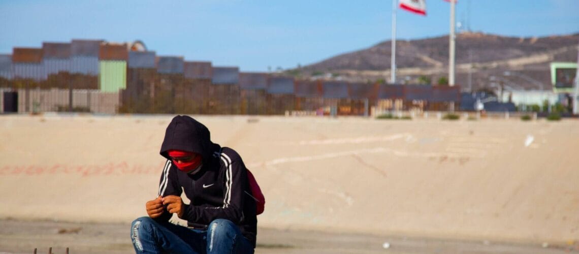 Este artículo habla sobre la expulsión de migrantes en la frontera. La imagen muestra a un hombre sentado con el muro fronterizo de fondo.
