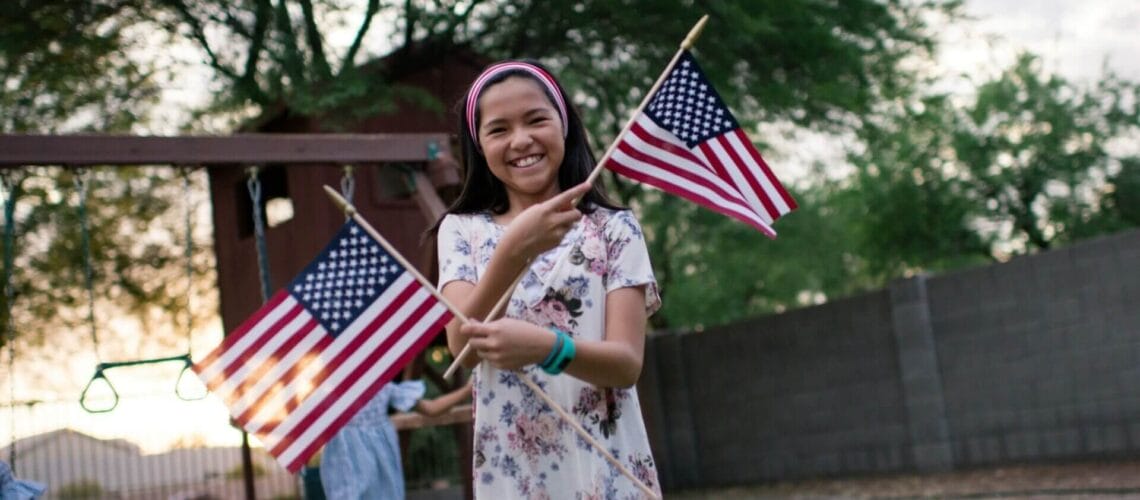 Este artículo habla sobre la ley "California ID's for All", que otorgará tarjetas de identificación estatal a residentes sin papeles. La imagen muestra una niña sonriendo. Sostiene dos banderines de Estados Unidos.